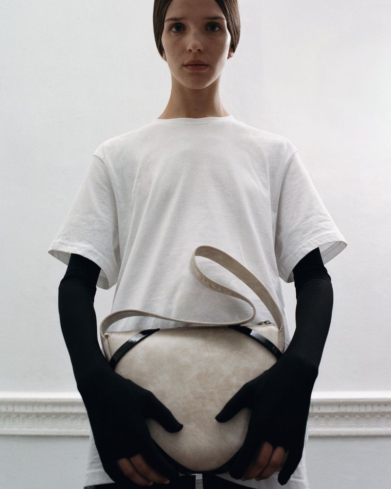 Fashion shot of a fair-skinned woman in a white t-shirt holding a neutral-toned bag in an indoor setting, minimalist studio lighting.