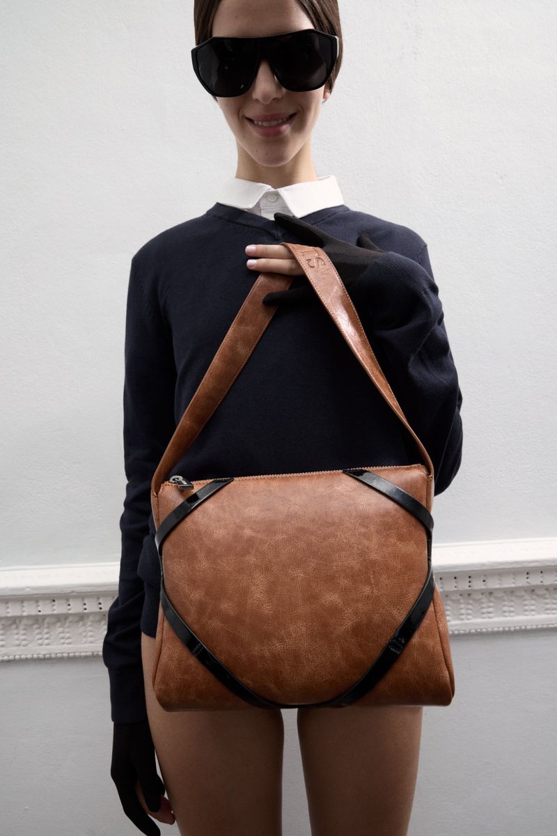 Brunette woman in sunglasses holding a brown leather crossbody bag in a fashion studio setting. Casual white shirt and black sweater.
