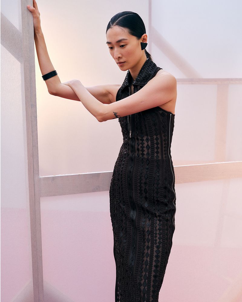 A slender Asian woman in a dark, patterned dress poses against a white and pink backdrop, highlighted by soft, diffused lighting, showcasing modern fashion.