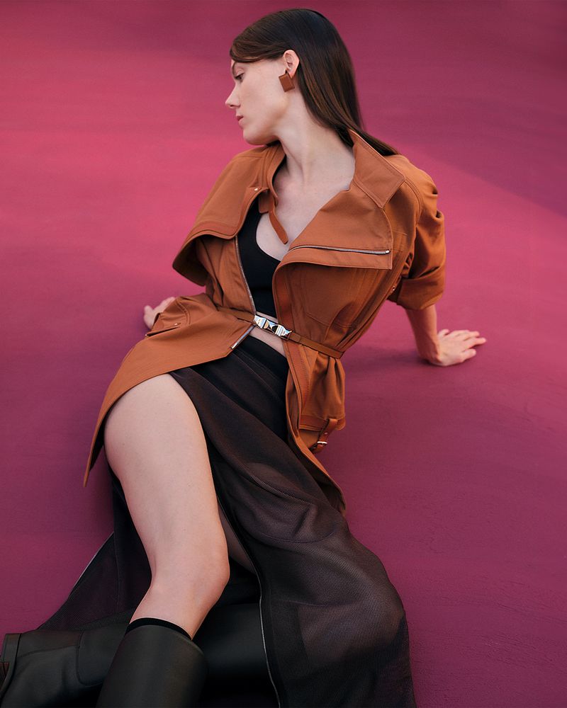 Elegant woman in a brown jacket and black skirt poses on a pink surface, showcasing fashion and style in a studio setting.