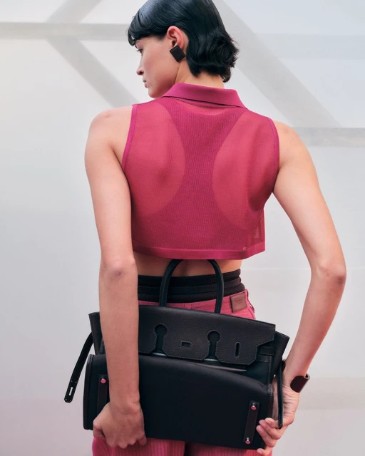 Back view of a model in a red mesh crop top and red pants holding a black leather handbag in an indoor studio setting.