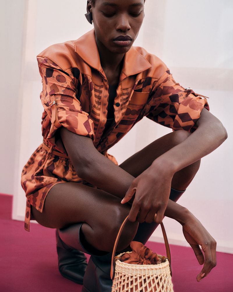 A stylish black woman in an orange patterned romper crouching indoors with a woven bag, showcasing a high-fashion editorial style.