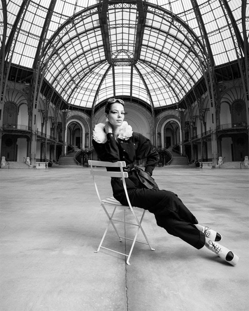 Black and white shot of a woman with dramatic eye makeup and aviator glasses sitting on a chair in a vast, ornate hall.