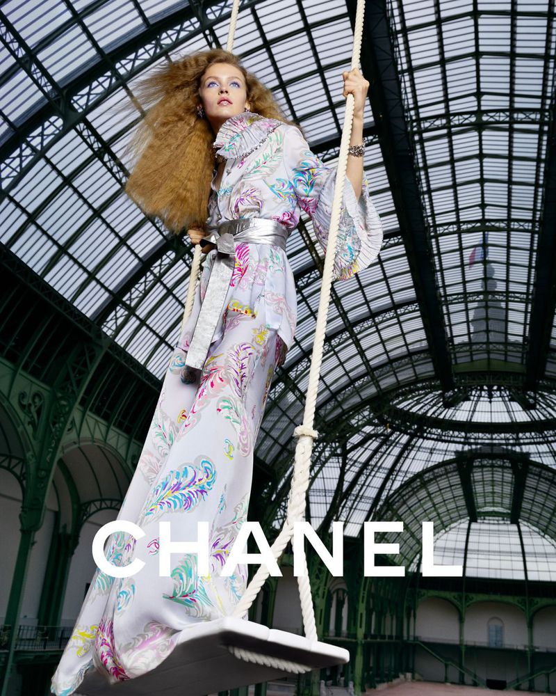 A fashion shot of a woman on a swing wearing a long floral dress, silver belt, and statement jewelry. She is in a building with an arched ceiling.