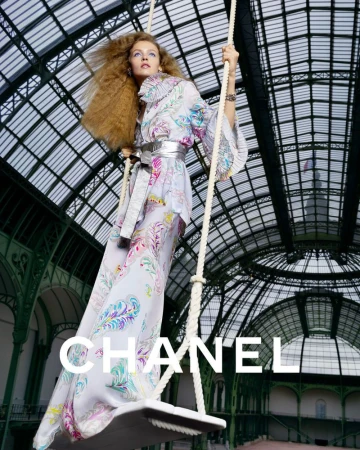 A fashion shot of a woman on a swing wearing a long floral dress, silver belt, and statement jewelry. She is in a building with an arched ceiling.