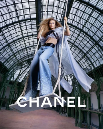 A blonde woman with long hair poses on a swing suspended in an indoor hall with glass ceilings, wearing a Chanel outfit and looking cool.