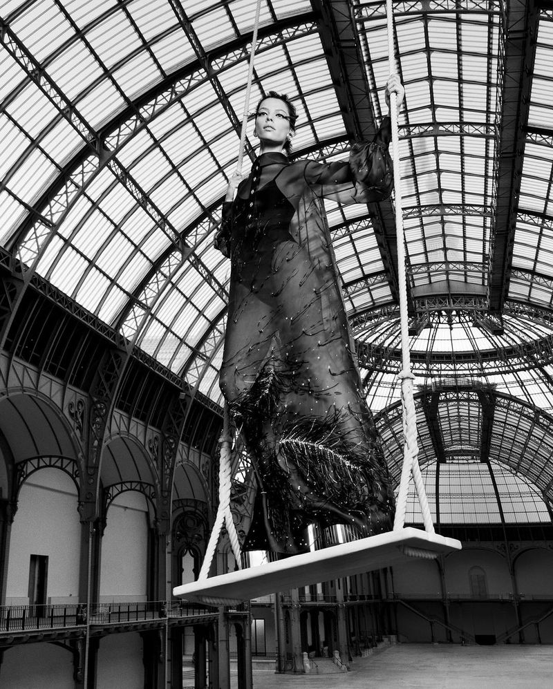 Black and white portrait of a woman in a sheer dress, posing on a swing inside a large, ornate hall with a high, arched ceiling.