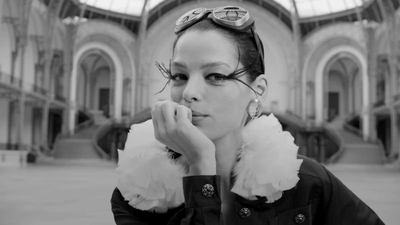 Black and white portrait of a woman in a jacket with a ruffled collar and goggles on her head inside a grand hall with arched openings.