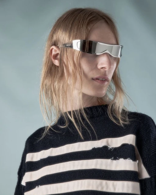 Close-up of a woman wearing reflective silver visor sunglasses and a striped sweater against a light blue background in a studio.