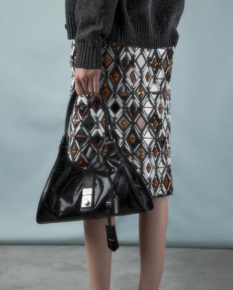 Close-up of a woman holding a Prada purse and wearing an embellished skirt and gray sweater, photographed in a photo studio.