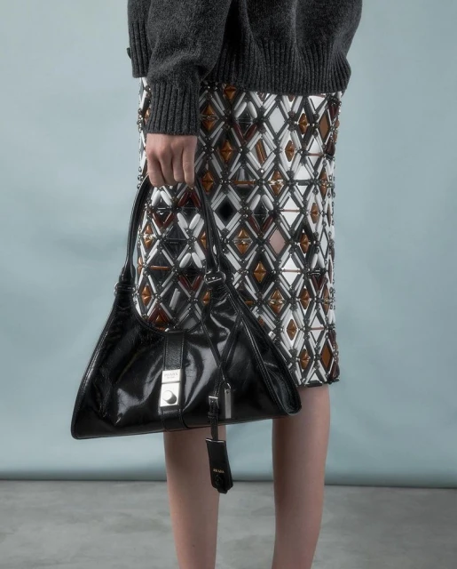 Close-up of a woman holding a Prada purse and wearing an embellished skirt and gray sweater, photographed in a photo studio.