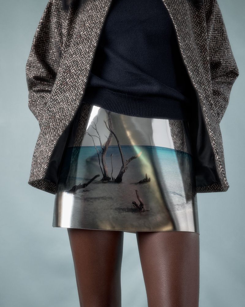Fashionable woman showcases a unique skirt with a nature scene, paired with a brown jacket and black sweater against a muted backdrop.
