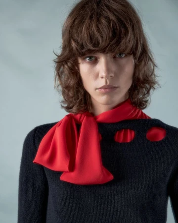 Model with brown hair wearing a black top and bright red scarf against a pale background