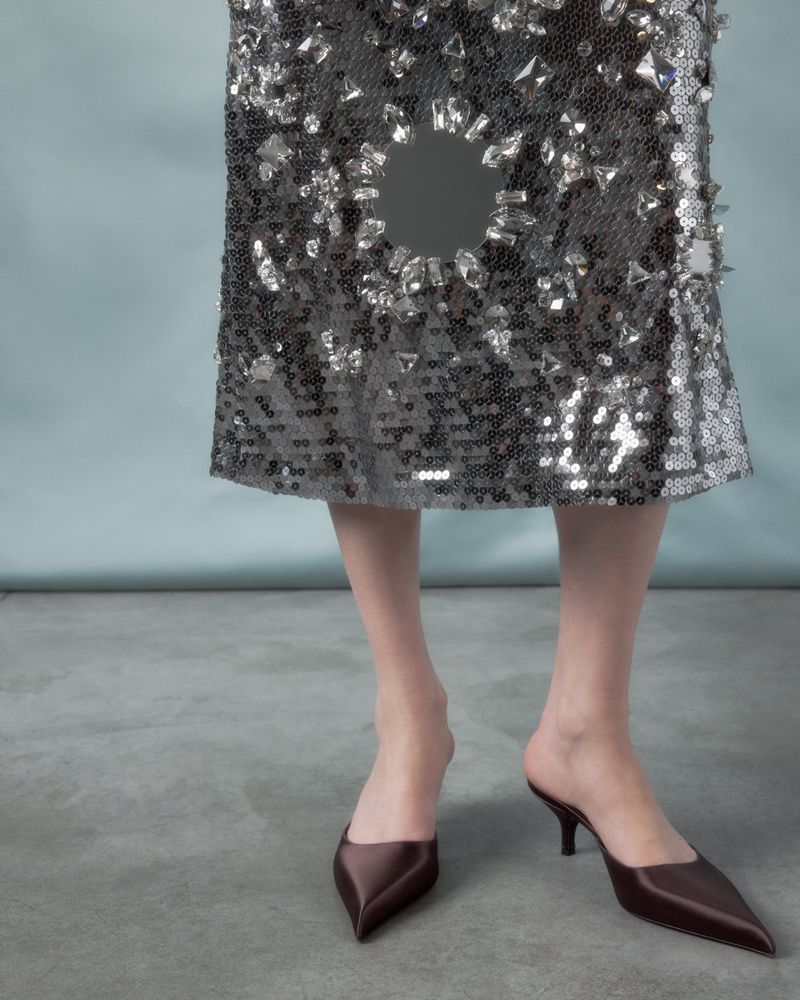 A model showcases a grey sequin skirt with a circular cutout, paired with brown pointed-toe heeled mules in a fashion studio.