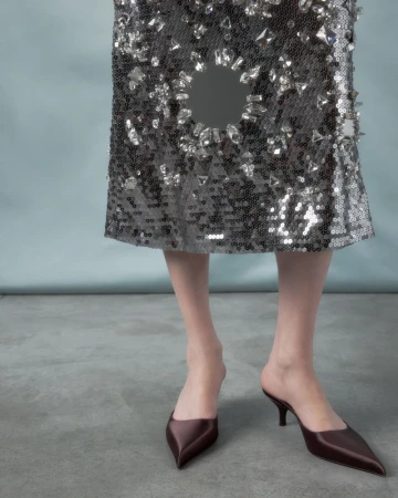 Close-up of a person wearing a crystal-embellished sequin skirt and pointed-toe kitten heel mules against a plain studio background.