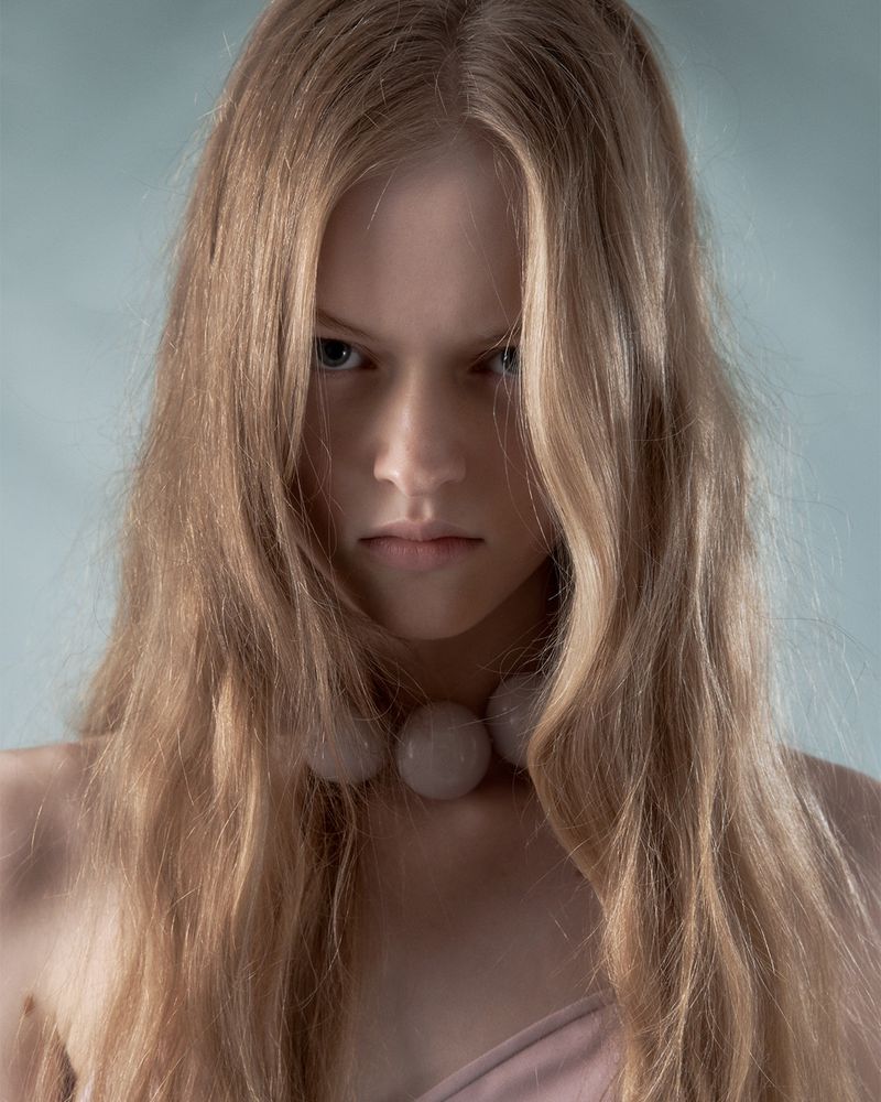Portrait of a fair-skinned young woman with long blonde hair, wearing a beaded necklace, looking intensely at the camera in a studio setting.