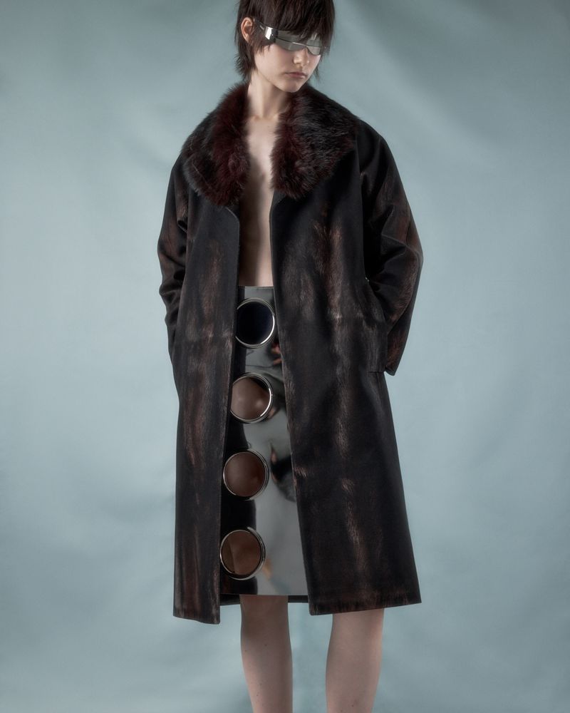 A young person poses in a long, dark brown coat with a fur collar and a silver skirt featuring cutout circle details against a blue background.