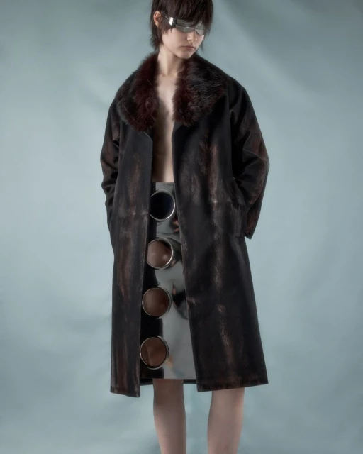 A young person poses in a long, dark brown coat with a fur collar and a silver skirt featuring cutout circle details against a blue background.
