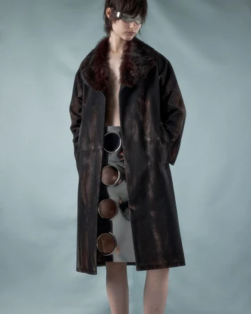 A young person poses in a long, dark brown coat with a fur collar and a silver skirt featuring cutout circle details against a blue background.