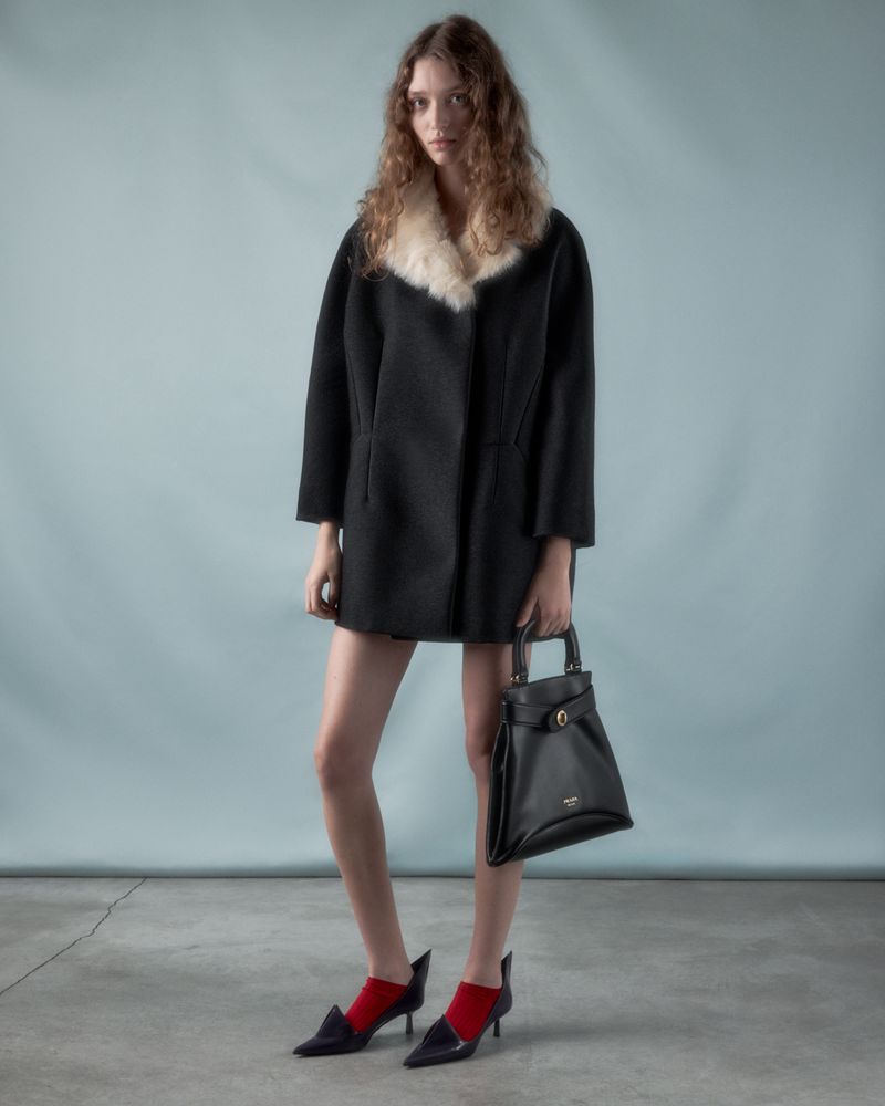 Model wearing a black coat with a white fur collar and holding a black handbag against a grey backdrop