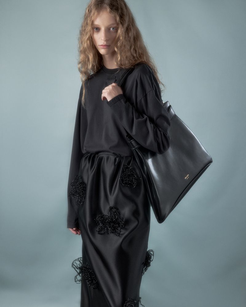 Fashion shot of a young adult woman posing in a black long-sleeve dress and holding a black leather tote bag on a blue background.