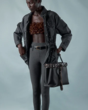 Dark-skinned woman poses in a studio, wearing a brown floral top, grey leggins, a black jacket, and a black studded purse.