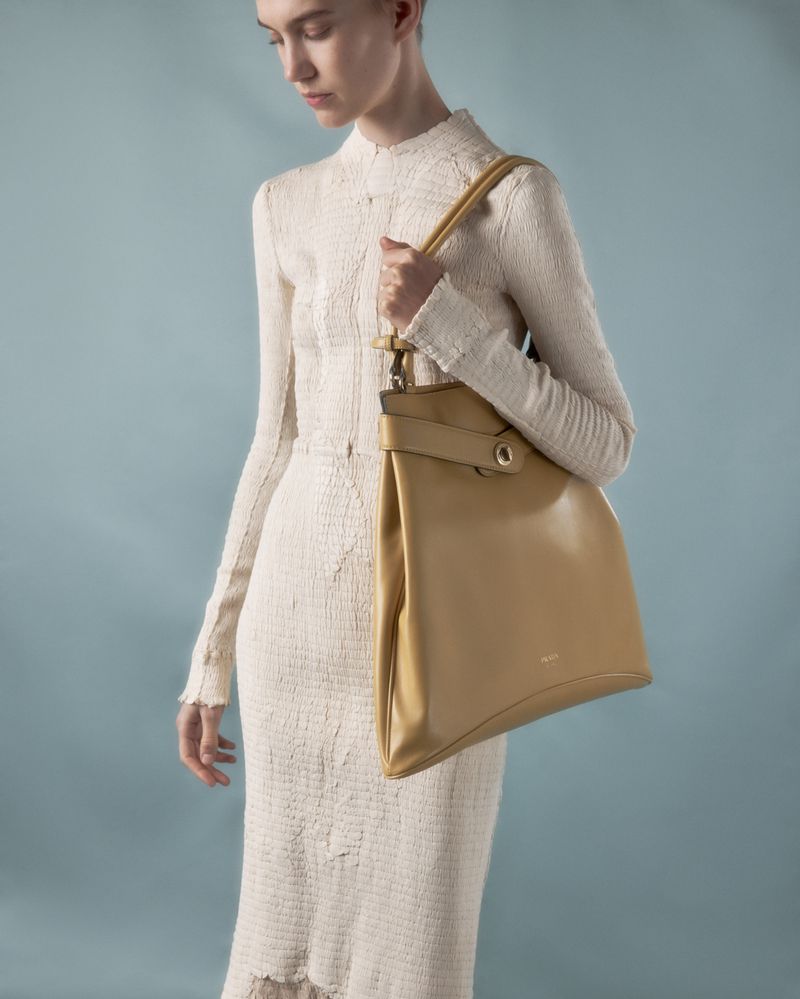 Model in cream textured dress holding a tan leather shoulder bag against a blue backdrop