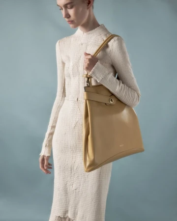 Model in cream textured dress holding a tan leather shoulder bag against a blue backdrop