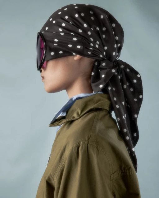 Woman in retro-futuristic fashion with polka dot headscarf and oversized goggles posing in profile against a light blue background.