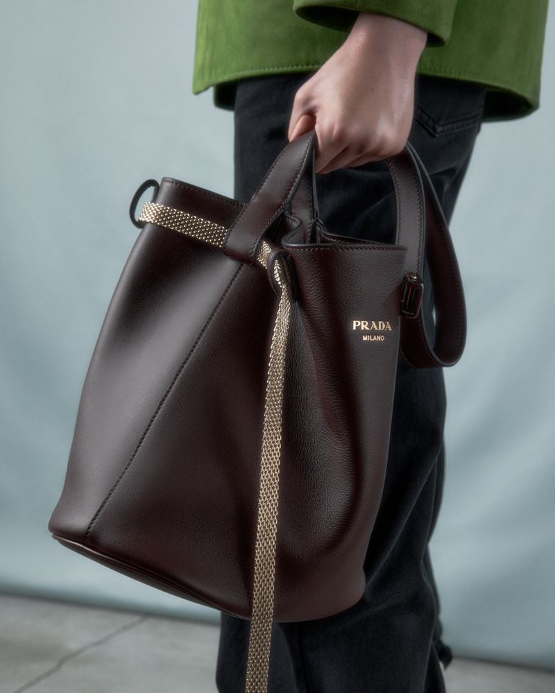 Leather Prada bucket bag with chain strap held by an adult with a green jacket and jeans against a light-blue wall in a studio.