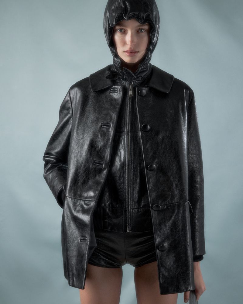 A woman poses in a shiny black hooded jacket and shorts against a light blue background, showcasing a modern, edgy fashion look.