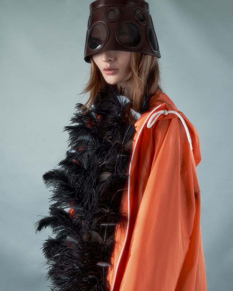 Eye-level shot of a fashion model wearing an orange hooded jacket and a brown leather head covering with black feather boa.