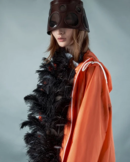 Eye-level shot of a fashion model wearing an orange hooded jacket and a brown leather head covering with black feather boa.
