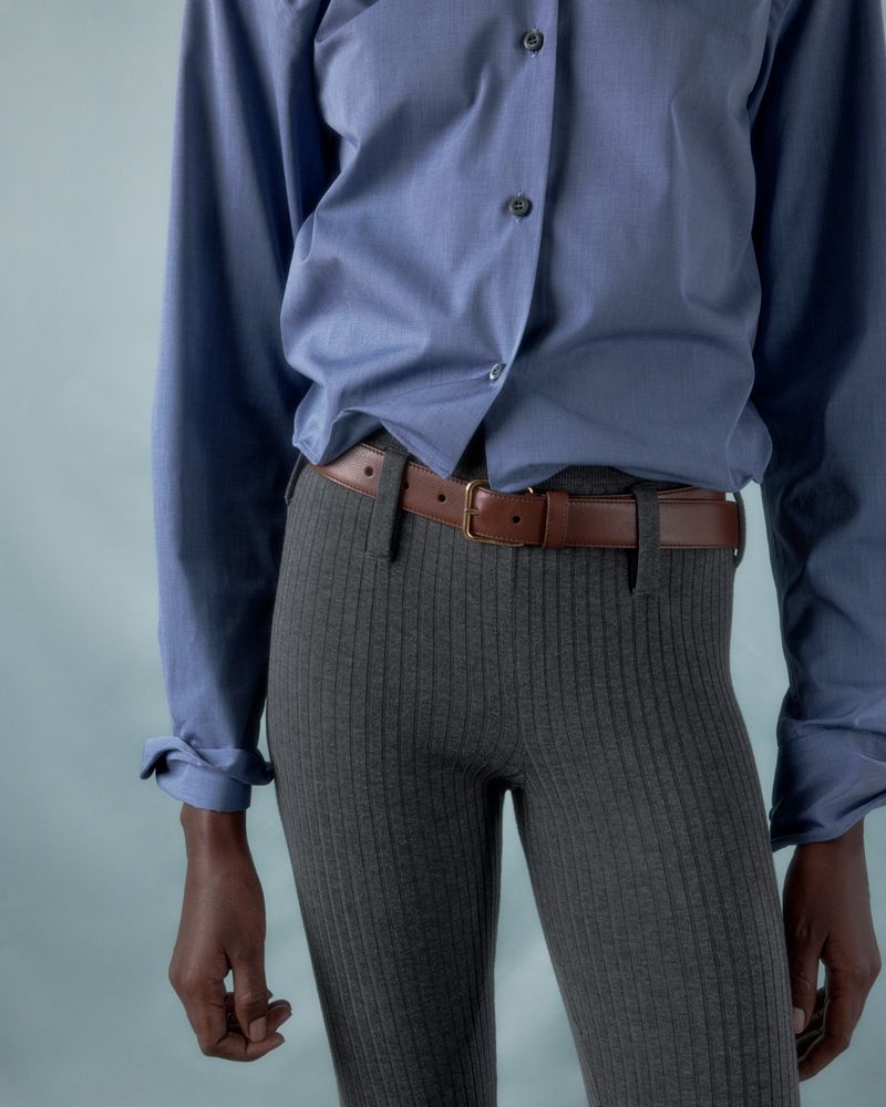 Person wearing a blue button-down shirt with a brown belt and gray ribbed pants against a light blue background in a studio setting.