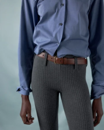 Person wearing a blue button-down shirt with a brown belt and gray ribbed pants against a light blue background in a studio setting.