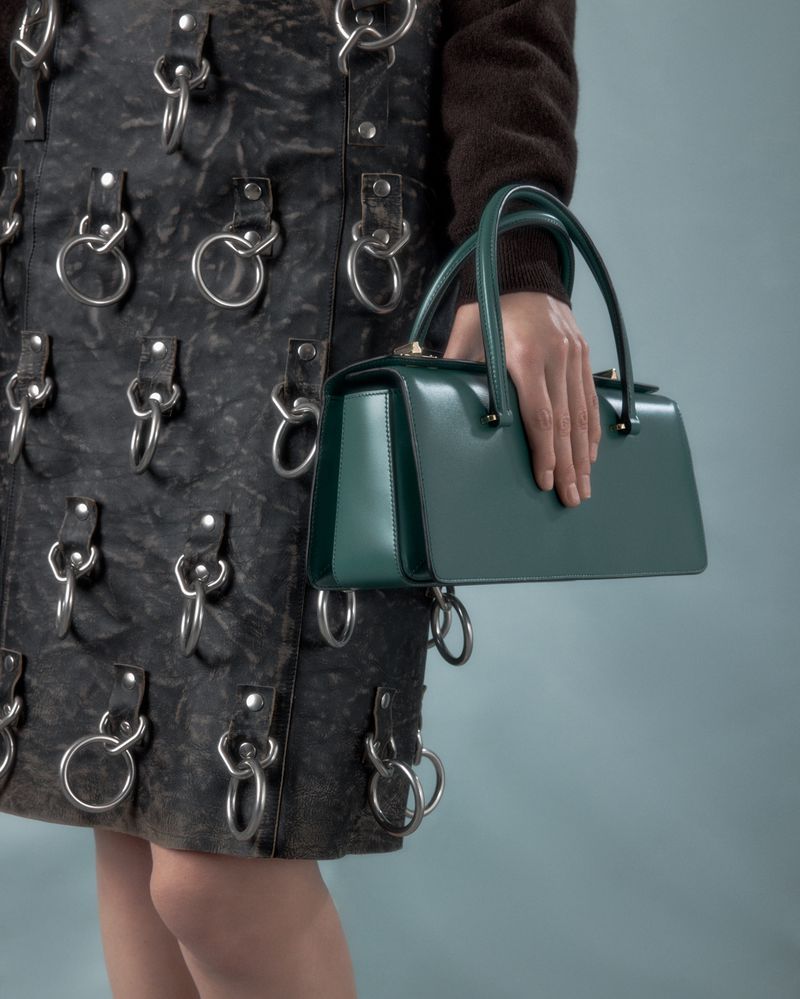Close-up of a woman's hand holding a structured green leather handbag with a black leather skirt featuring metal ring attachments.