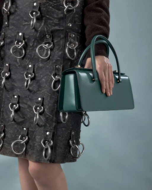 Close-up of a woman's hand holding a structured green leather handbag with a black leather skirt featuring metal ring attachments.