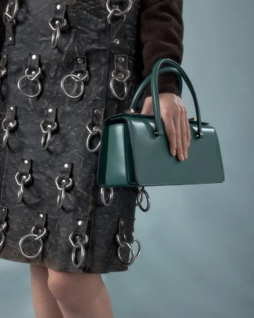 Close-up of a woman's hand holding a structured green leather handbag with a black leather skirt featuring metal ring attachments.