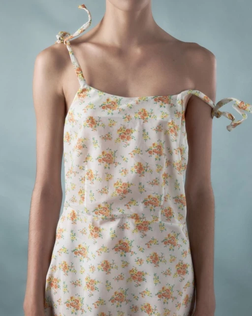 Woman in a white dress with a floral print featuring orange flowers and green leaves, tied at the shoulders, standing against a blue background.