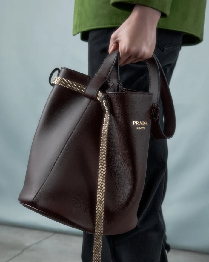 Model holding a dark brown leather Prada bucket bag with gold lettering against a plain studio background.