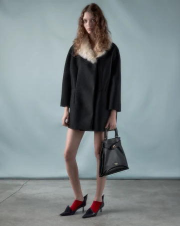 Model wearing a black coat with a white fur collar and holding a black handbag against a grey backdrop