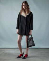 Model wearing a black coat with a white fur collar and holding a black handbag against a grey backdrop