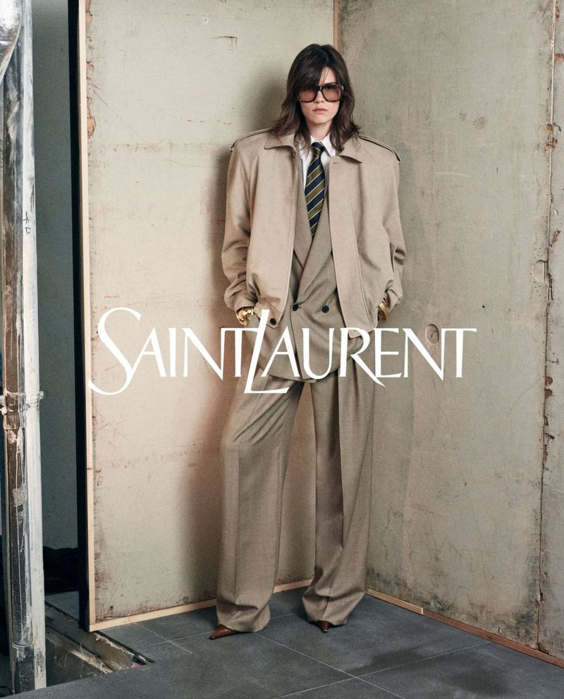 An adult model poses in a light-tan three-piece suit and matching trench coat in a studio, with a Saint Laurent logo visible in the frame.