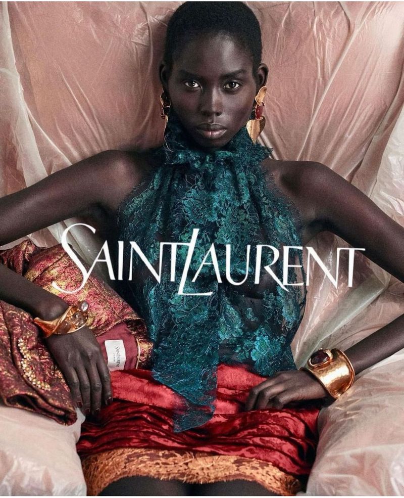 Fashion image featuring a Black woman model in a teal top and red skirt, wearing gold bracelets, posing in front of a light background.