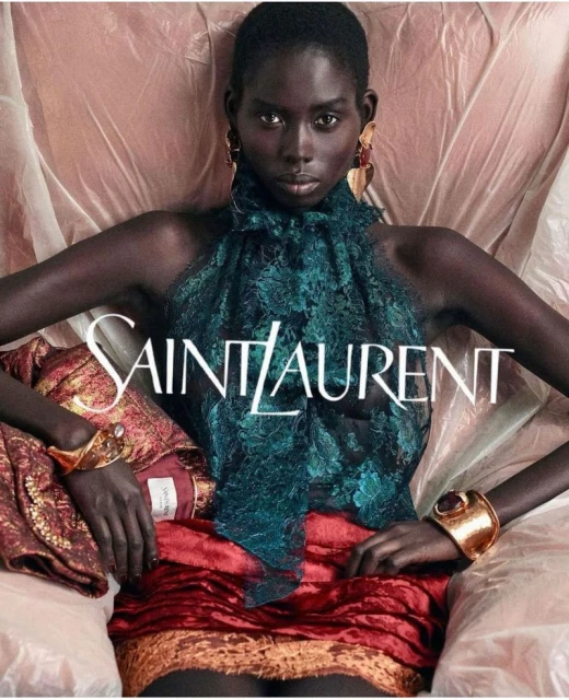 Fashion image featuring a Black woman model in a teal top and red skirt, wearing gold bracelets, posing in front of a light background.