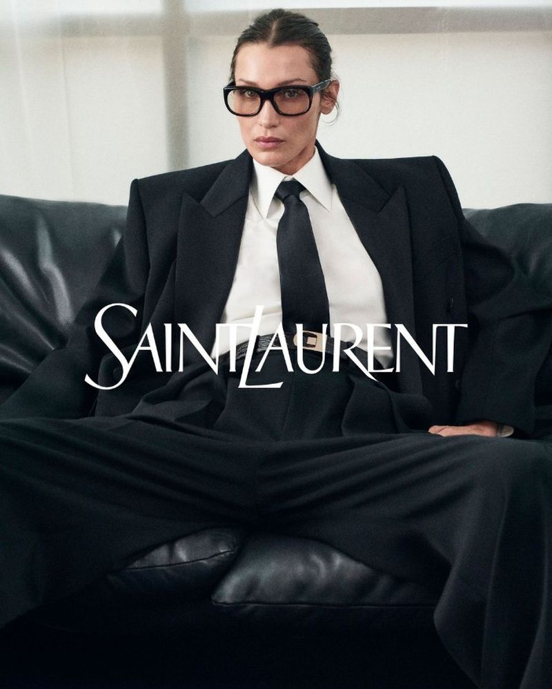 Bella Hadid fashion portrait: woman in a black suit and glasses sits on a black leather couch in a studio setting, Saint Laurent logo visible.