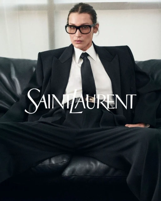 Bella Hadid fashion portrait: woman in a black suit and glasses sits on a black leather couch in a studio setting, Saint Laurent logo visible.