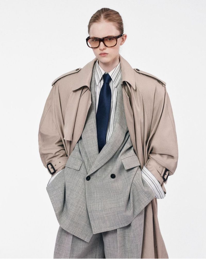 Fashionable young woman wearing oversized blazer and trench coat posing in a photo studio against a clean white backdrop.