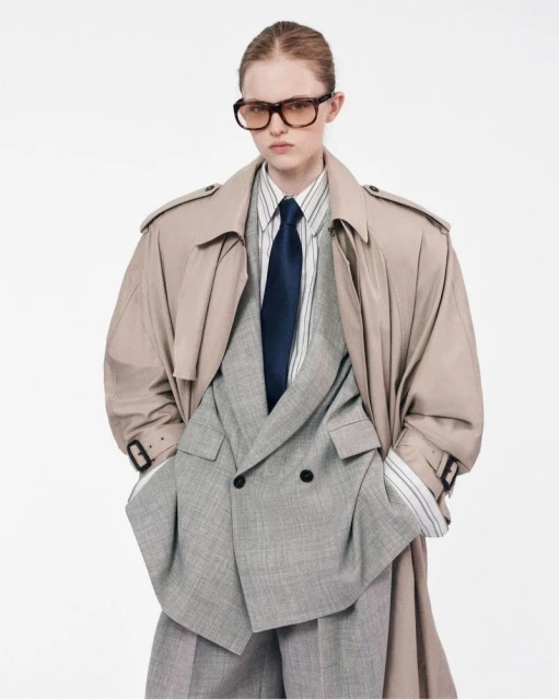 Fashionable young woman wearing oversized blazer and trench coat posing in a photo studio against a clean white backdrop.