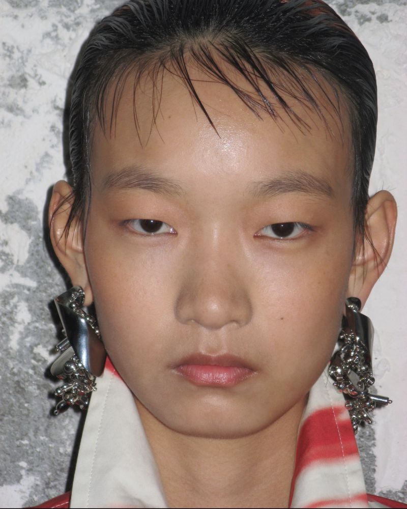 Close-up portrait of a young Asian woman with slicked-back hair, wearing statement silver earrings, against a textured grey backdrop.